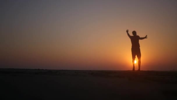 Een man loopt langs het strand en draait met zijn handen omhoog in de zonsondergang. HD, 1920 x 1080. Slow motion. — Stockvideo