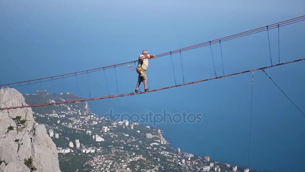 Ein Mann läuft auf einer Hängeseilbrücke in großer Höhe vor dem Hintergrund von Felsen, Meer und Stadt. hd, 1920x1080. Zeitlupe. — Stockvideo