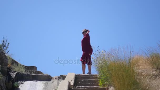 La ragazza sta girando, in piedi sulla cima delle scale, in un flusso di vento contro il cielo e l'erba. 3840x2160, 4K — Video Stock