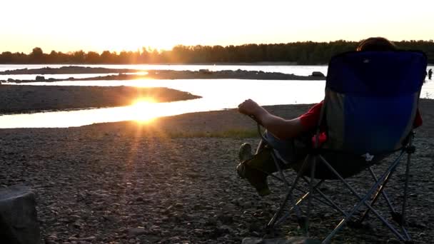 Genießen Sie den Sonnenuntergang auf einem Hocker auf einer steinigen Bank in der Nähe des Flusses bei Sonnenuntergang. Nahaufnahme. Zeitlupe, 1920x1080, hd — Stockvideo