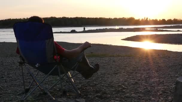 Un homme ne se repose pas sur la plage dans une chaise pliante, jette son pied sur sa jambe, profite du coucher de soleil. au ralenti, 1920x1080, en haute qualité — Video