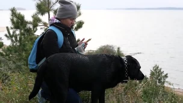 一个小女孩在一顶帽子的裤子和一条大狗在她的手，在湖的高高的河岸上坐着电话。1920 x 1080 高清，慢动作 — 图库视频影像