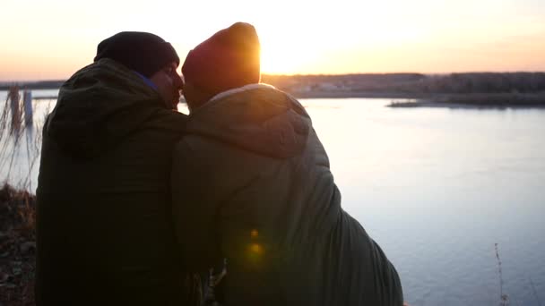 Un couple aimant sur la rive de la rivière en vestes et casquettes est assis sur un fond de coucher de soleil avec un effet de lentille. ralenti, 1920x1080, full hd — Video