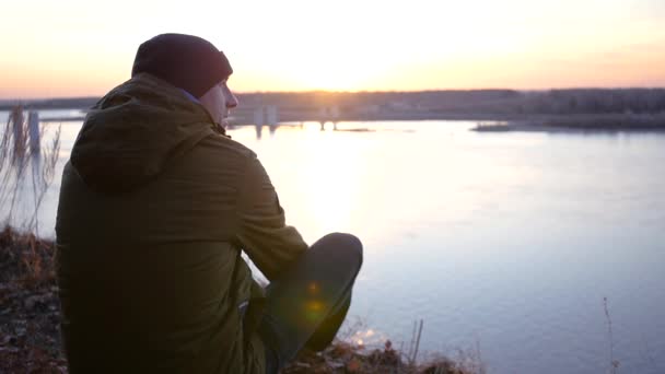 Um jovem de casaco e chapéu senta-se na margem do rio perto da ponte e admira o belo pôr-do-sol. câmera lenta, 1920x1080, hd completo — Vídeo de Stock