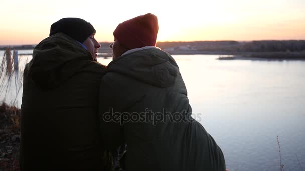 Δύο άνθρωποι κάθονται στην όχθη του ποταμού, κρυφοκοιτάζουν και φιλιά με φόντο ένα μαγευτικό ηλιοβασίλεμα στη γέφυρα. αργή κίνηση, 1920 x 1080, full hd — Αρχείο Βίντεο