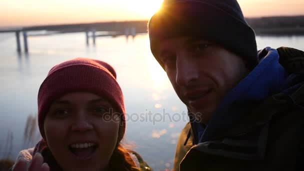 Ungt par, man och kvinna tar ett foto på telefonen, skrattar åt kameran på en solnedgång bakgrund vid floden. Slowmotion, 1920 x 1080, full hd — Stockvideo