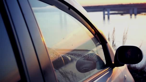 El hombre abre una ventana en el coche y admira la hermosa puesta de sol en el río en el puente, sentado en el estacionamiento en el coche. cámara lenta, 1920x1080, full hd — Vídeos de Stock