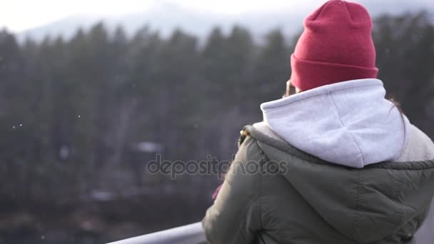 Une jeune fille se tient dans une veste et un chapeau sur le pont et prend une photo, lors d'une chute de neige. ralenti, 1920x1080, full hd — Video