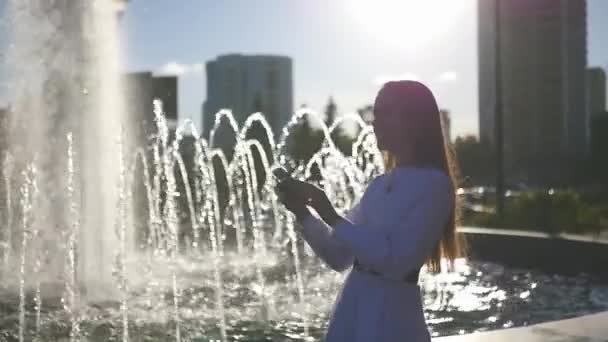 Een mooi meisje staat in de buurt van een zomer-fontein en maakt een selfie. HD, 1920 x 1080. Slow motion. — Stockvideo