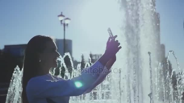 魅力的な女の子は、市内の泉のそばに立って、スマート フォンで selfie を行います。1920 x 1080 の Hdスローモーション. — ストック動画
