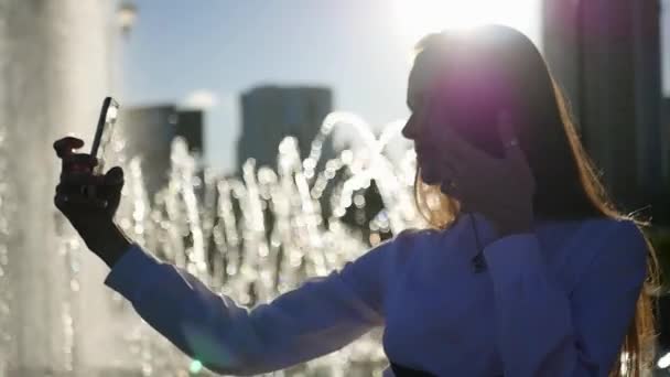 Красива дівчина з довгим волоссям позує на камеру, робить фото, що стоїть біля фонтану з розмитими променями сонця. HD, 1920x1080. повільний рух . — стокове відео