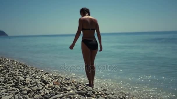 Une fille mince en maillot de bain, marchant le long de la plage avec des cailloux près de la mer bleue. HD, 1920 x 1080. au ralenti . — Video