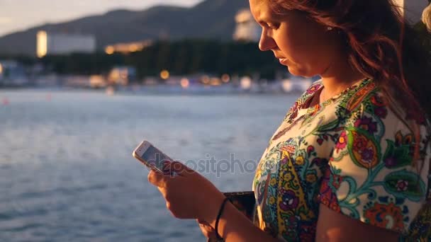 La chica está de pie en el paseo marítimo junto al mar, escribiendo algo en el teléfono inteligente al atardecer. HD, 1920x1080. cámara lenta . — Vídeos de Stock