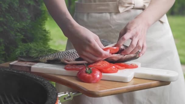 La casalinga mette pomodori a fette nel pesce crudo prima di cuocerlo, pesce carpa sulla tavola, cucinare pasti di pesce all'aperto, frutti di mare alla griglia, barbecue e fuoco. — Video Stock