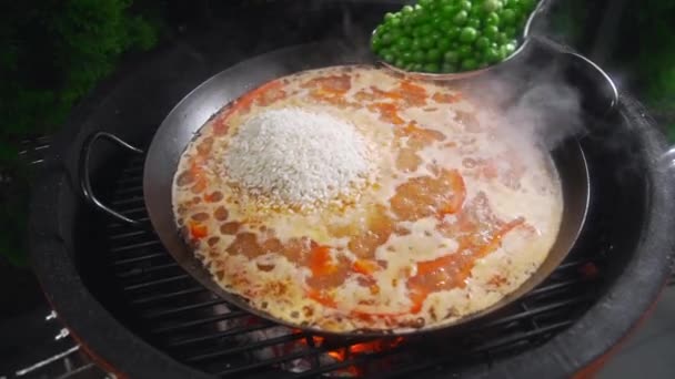 A szakács a kandalló teszi a paella és zöld borsó hozzáadja az egyéb összetevők, a nagy forró serpenyőben, grill és grill, a spanyol étel, a paella, tenger gyümölcsei, étel, főzés szabadban — Stock videók