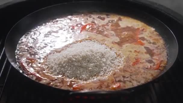 O cozinheiro faz paella na lareira e adiciona arroz aos outros ingredientes na panela quente grande, churrasqueira e churrasqueira, comida espanhola, paella com frutos do mar, comida cozinhando ao ar livre — Vídeo de Stock