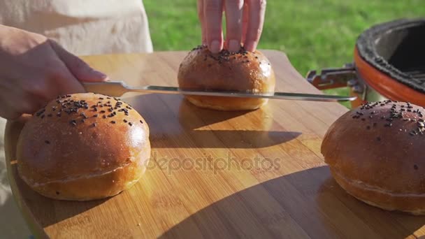 Femme au foyer coupe le pain en deux par couteau sur la planche en bois pour hamburger grill faire à l'extérieur — Video