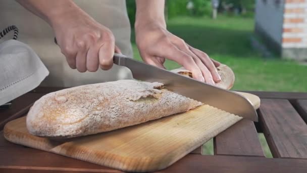 Ama de casa corta la baguette por la mitad por el cuchillo en el tablero de madera al aire libre, harina y panadería, haciendo las arenas brujas — Vídeo de stock