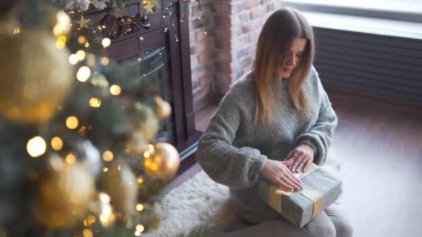 Mujer bonita en suéter gris decora la caja de regalos y lo pone bajo el árbol de Navidad, árbol de Navidad brillante, guirnaldas y bolas, regalos de año nuevo, preparaciones de Navidad, víspera de año nuevo — Vídeo de stock