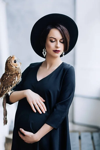 Femeie gravidă caucaziană cu machiaj în rochie neagră și pălărie neagră cu bufniță așezată pe braț, portretul mamei viitoare, sarcină fericită, portret de modă, femeie în negru — Fotografie, imagine de stoc
