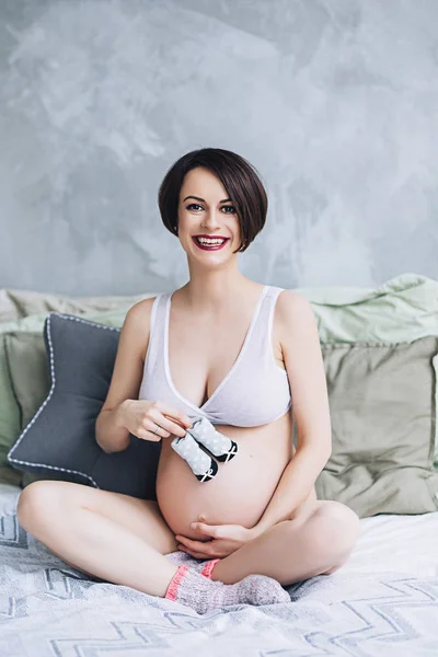 Fericit frumoasă femeie caucaziană însărcinată cu burtă mare în lenjerie albă ține copii rochie, femeie stă pe pat în dormitor, viitoarele chirie, așteptând un copil, maternitate și îngrijire — Fotografie, imagine de stoc