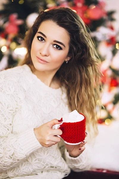 Femeia stă lângă pomul de Crăciun cu o ceașcă de ciocolată caldă, o femeie frumoasă în pulover, atmosferă de vacanță și ghirlande, o femeie albă frumoasă — Fotografie, imagine de stoc