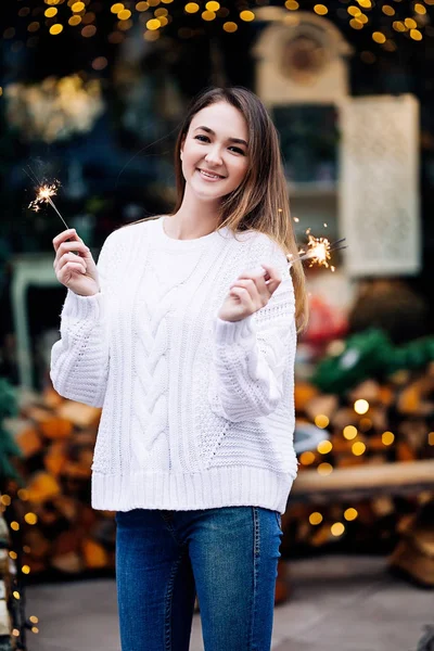 Happy white girl with long dark hair in sweater, garlands in bokeh, holiday cozy atmosphere, smiling woman with bengal lights, outdoors portrait — Stock Photo, Image