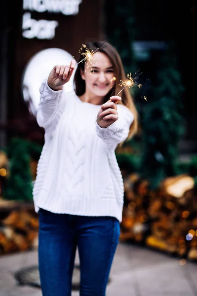 Happy white girl with long dark hair in sweater, garlands in bokeh, holiday cozy atmosphere, smiling woman with bengal lights, outdoors portrait — Stock Photo, Image