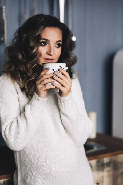 Caucasian pregnant woman with make up and curly hair in warm white sweater holds cup with hot chocolate, portrait of future mother, happy pregnancy, fashion portrait, stylish pregnancy