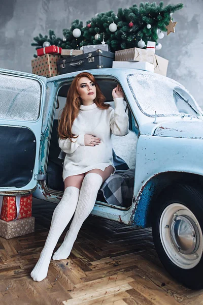 Frumoasă femeie gravidă elegantă în pulover lung alb cu păr lung roșu stă în mașina deschisă elegantă mică, cu brad de Crăciun și cadouri și își ține burta, frumusețea sarcinii, așteptând o — Fotografie, imagine de stoc