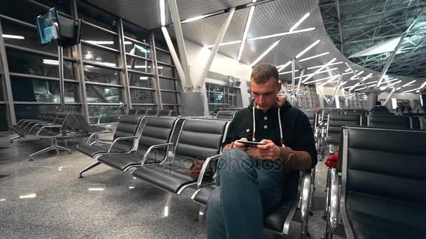 旅行者は空港の待っているホールで彼の飛行を待機する、空気、携帯電話の画面に男の視線は、待っているホールで孤独な乗客によって男の旅 — ストック動画