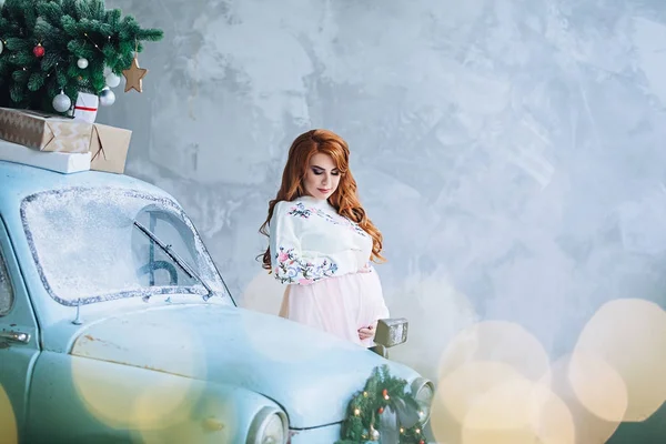 Frumoasă femeie gravidă elegantă în pulover lung alb cu păr lung roșu stă aproape de mașina mică elegantă cu brad de Crăciun și prezintă și ține burta, frumusețea sarcinii, așteptând un copil — Fotografie, imagine de stoc