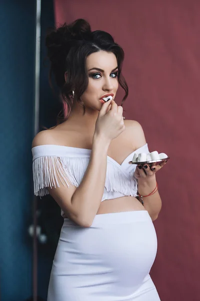 Model hamil yang cantik dengan rambut coklat panjang dengan gaun putih tersenyum dan makan marshmallow, potret wanita hamil yang cantik, perut hamil yang lucu, ibu masa depan — Stok Foto