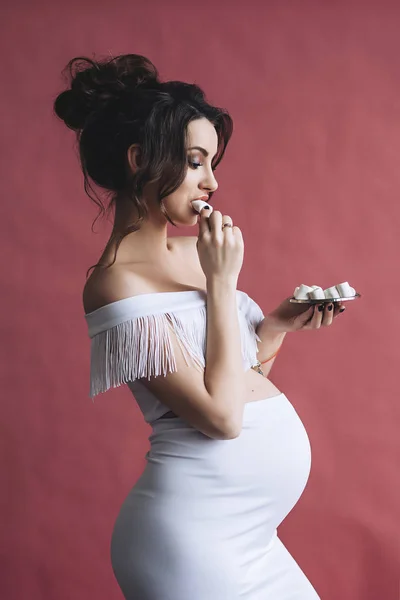 Modelo grávida bonita com cabelos castanhos longos no vestido branco sorri e come marshmallows, retrato de mulher grávida bonita, barriga grávida bonito, futura mãe — Fotografia de Stock