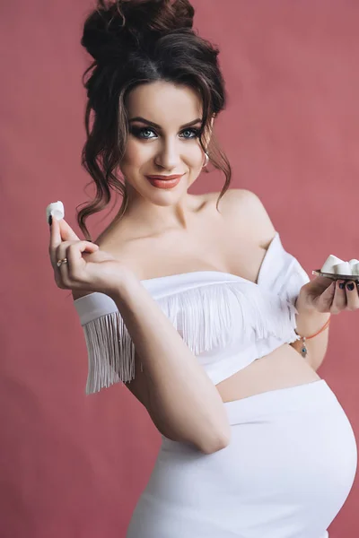 Modelo grávida bonita com cabelos castanhos longos no vestido branco sorri e come marshmallows, retrato de mulher grávida bonita, barriga grávida bonito, futura mãe — Fotografia de Stock