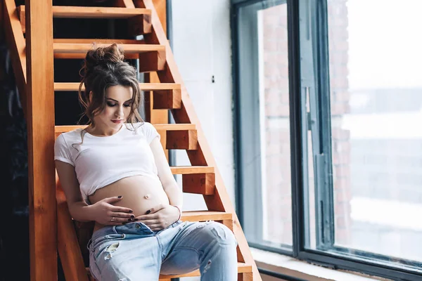 Beyaz t-shirt ve kot uzun kahverengi saçlı güzel şımarık hamile model ahşap merdivenlerden oturur ve pencerenin güzel hamile kadın, sevimli karın portresi görünüyor