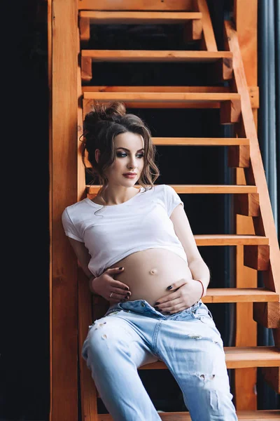 Modelo grávida atrevido bonito com cabelos castanhos longos na camiseta branca e jeans azul senta-se nas escadas de madeira e olha para a janela, retrato de mulher grávida bonita, barriga grávida bonito — Fotografia de Stock