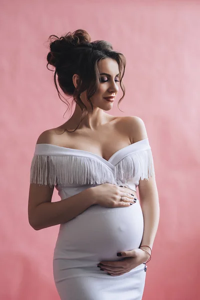 Bardzo piękne kaukaski kobieta w ciąży z tworzą i ciemne włosy w białej sukni posiada jej brzuch, portret przyszłej matki, szczęśliwy, ciąża, moda piękny portret stylowy ciąży — Zdjęcie stockowe