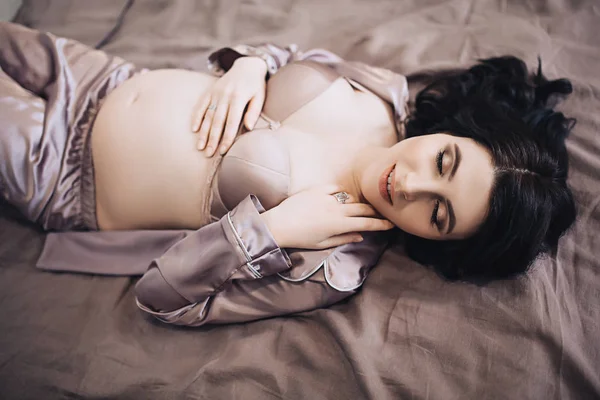 Krásná stylová těhotná žena s dlouhými tmavými vlasy drží její břicho a lehne na postel v pyžama, krása těhotenství, čeká dítě, budoucí rodiče, stát se matkou, nový život — Stock fotografie
