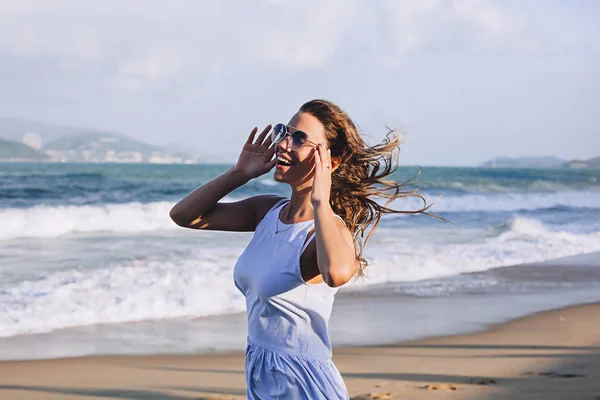 Vacker kvinna kort sarafan och sol glasögon med fladdrande hår står på stranden nära havet och vågorna, Sexig tjej på havet, vacker kvinna på semester — Stockfoto
