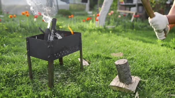 Mannen chopps trä av yxan nära brasan, Bbq på bakgården, att göra glödande kol för matlagning på den öppna elden, sommar barbecue — Stockvideo