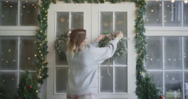 Mujer en suéter caliente lanza decorar las puertas principales con corona de Navidad, la decoración de la casa de Navidad, acogedor ambiente nuevo hogar año, 4k DCI 120fps Prores HQ — Vídeo de stock