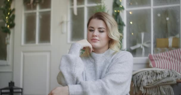 Ευρωπαϊκή νεαρή ελκυστική γυναίκα σε ζεστό πουλόβερ κάθεται στο εσωτερικό σκανδιναβικό και κοιτάζει προς την κάμερα, ζεστή ατμόσφαιρα σπίτι Χριστούγεννα και τα νέα χρόνια διακοσμήσεις, 4k DCI 120fps Prores Hq — Αρχείο Βίντεο