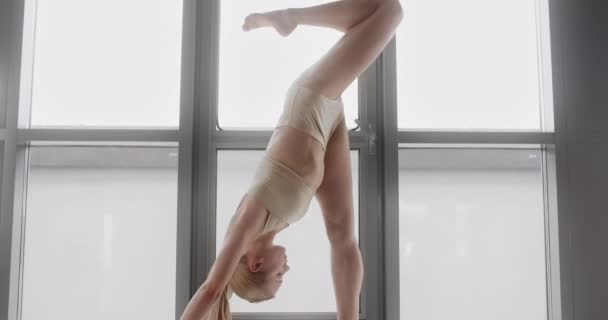 Atractiva joven rubia hace trucos gimnásticos acrobáticos en cámara lenta en el alféizar de la ventana del estudio de yoga, mujer flexible hace ejercicios acrobáticos y plásticos, yoga y estiramiento, 4k DCI 60p — Vídeo de stock