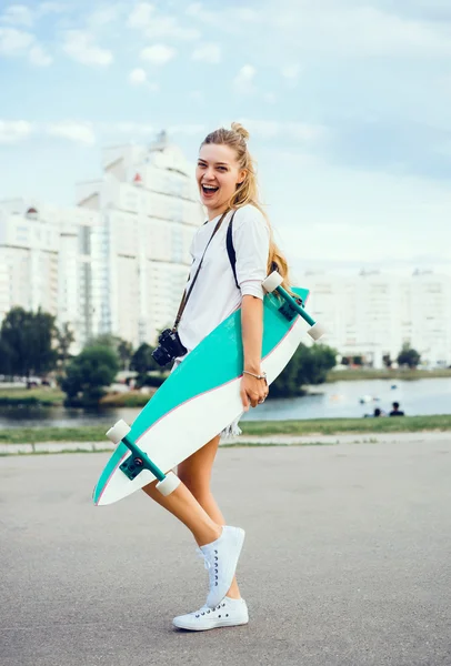 Молодая женщина позирует на улице с длинной доской, скейтборд, хипстерский стиль, открытый портрет крупным планом, кроссовки, джинсы джинсы — стоковое фото