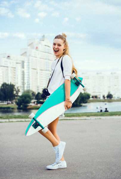 молодая женщина позирует на улице с длинной доской, скейтборд, хипстерский стиль, открытый портрет крупным планом, кроссовки, джинсы джинсы
