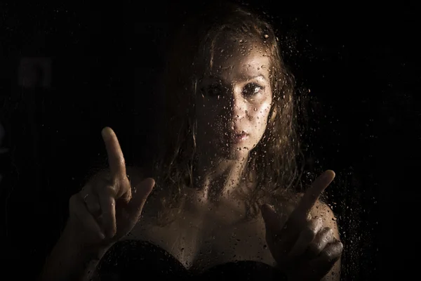 sexy young woman, posing behind transparent glass covered by water drops. melancholy and sad female portrait