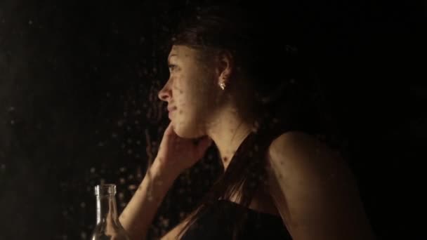 Молода сумна жінка за вікном з краплями на ньому. самотня дівчина п'є бренді з пляшки і плаче — стокове відео