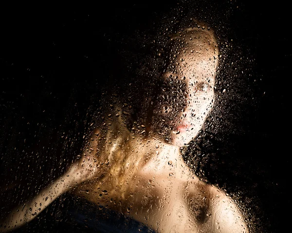 Sexy joven, posando detrás de vidrio transparente cubierto por gotas de agua. retrato femenino melancólico y triste — Foto de Stock