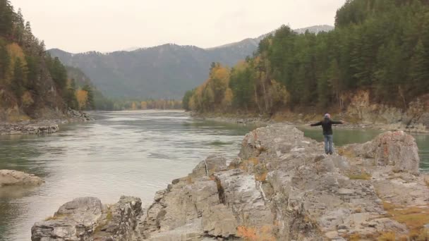 Berg rivier stroomt tussen rotsen met pijnbomen. reiziger staat op een grote rots aan de rivier — Stockvideo